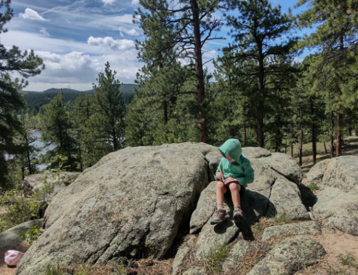 Little boy in green hooded sweatshirt sitting on a rock writing in a notebook.