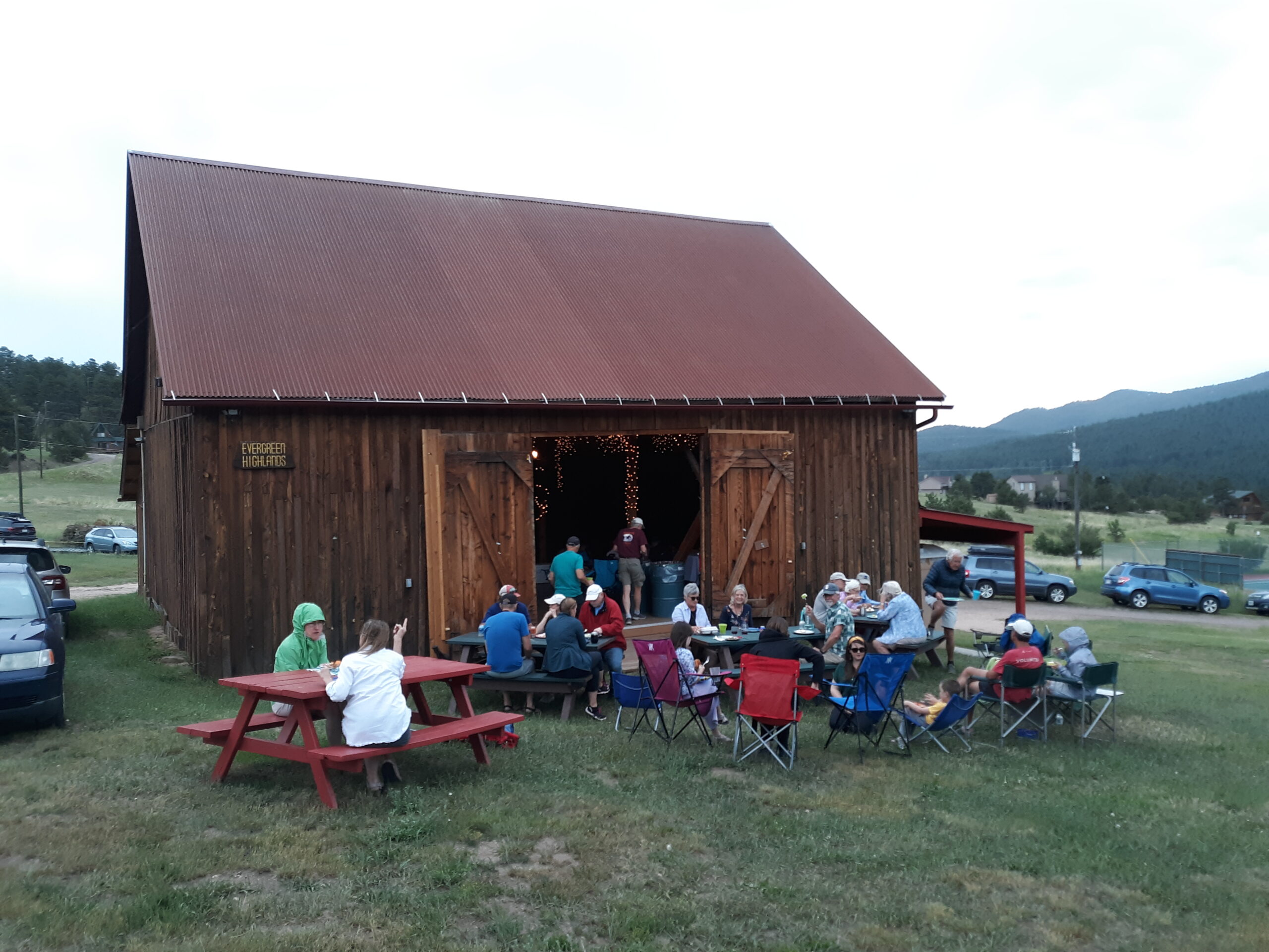 Several brightly dressed people sitting at picnic tables and on camp chairs in front of a rustic barn that has it's doors wide open. It looks like its about to pour rain.