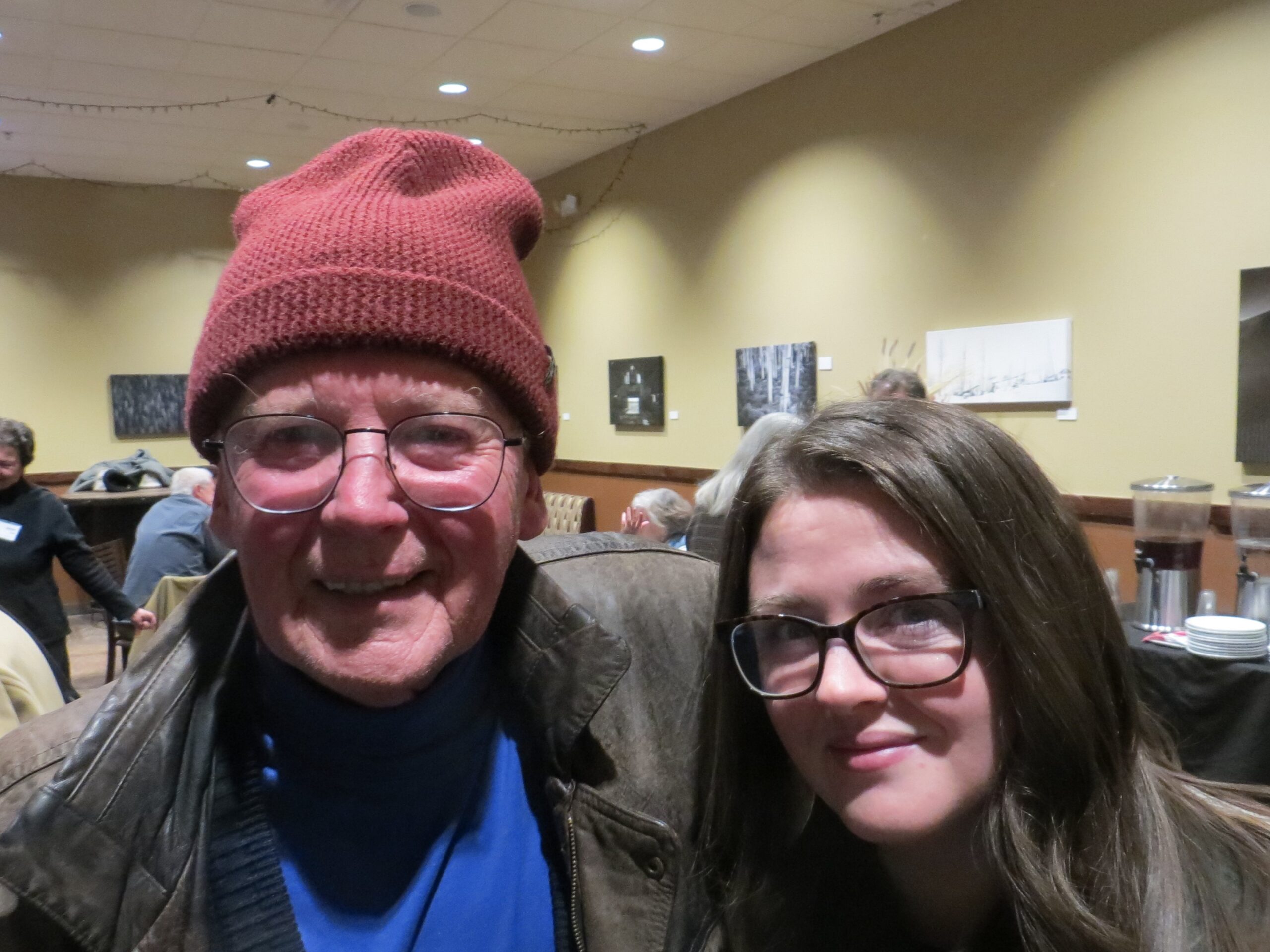 Close up faces of a man wearing wire rimmed classes, a brick red colored beanie, a blue sweater and a brown leather jacket and a women with dark framed glasses and long dark hair. Both are smiling sweetly.