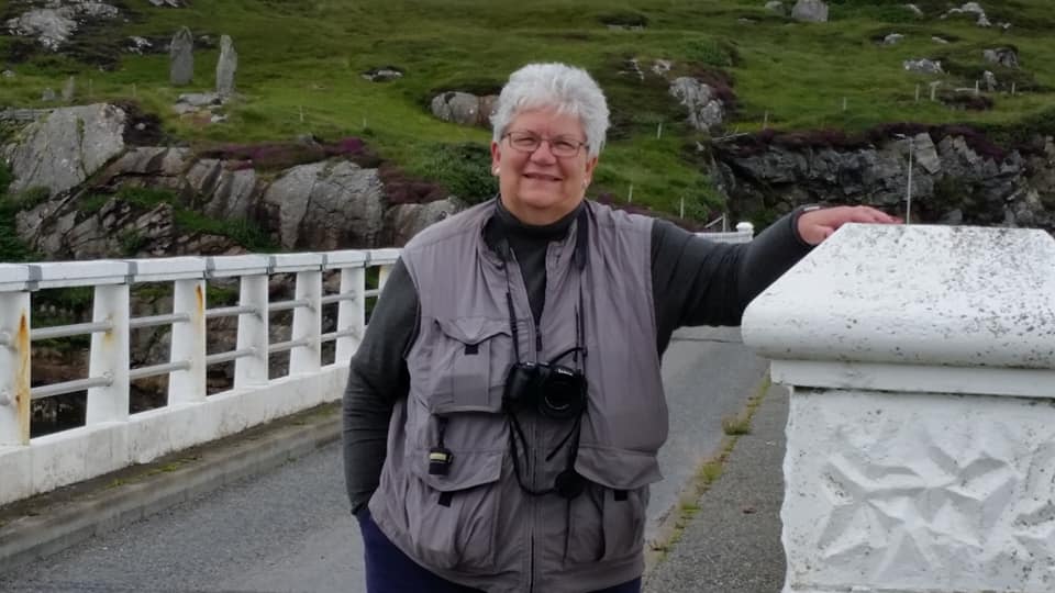 A grey haired woman, earing a black turtleneck, a grey travel vest and a camera around her neck, standing on a bridge. The background is a lush green and rocky landscape - England, maybe? Or Scotland?