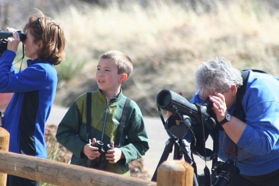 A woman wearing a blue jacket, looking through binoculars; a young boy in a green jacket holding binoculars, and a woman in a blue jacket, looking through a spotting scope.