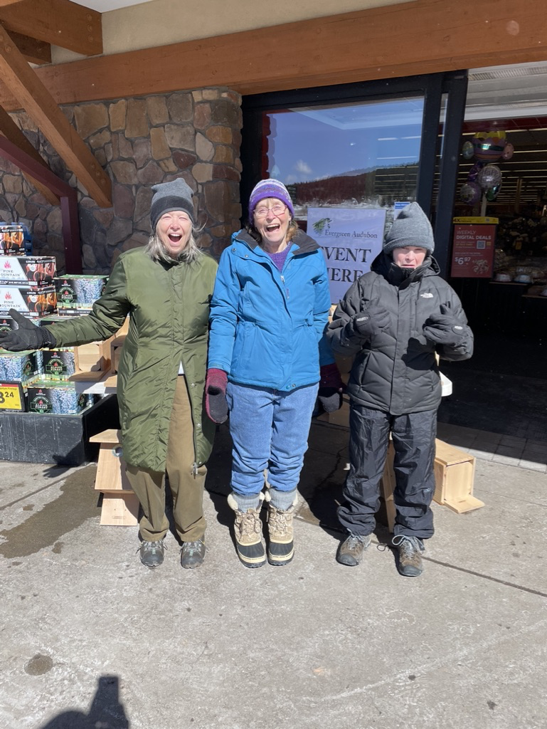 Three adult women in winter clothing in front of a table of nest boxes (bird houses)