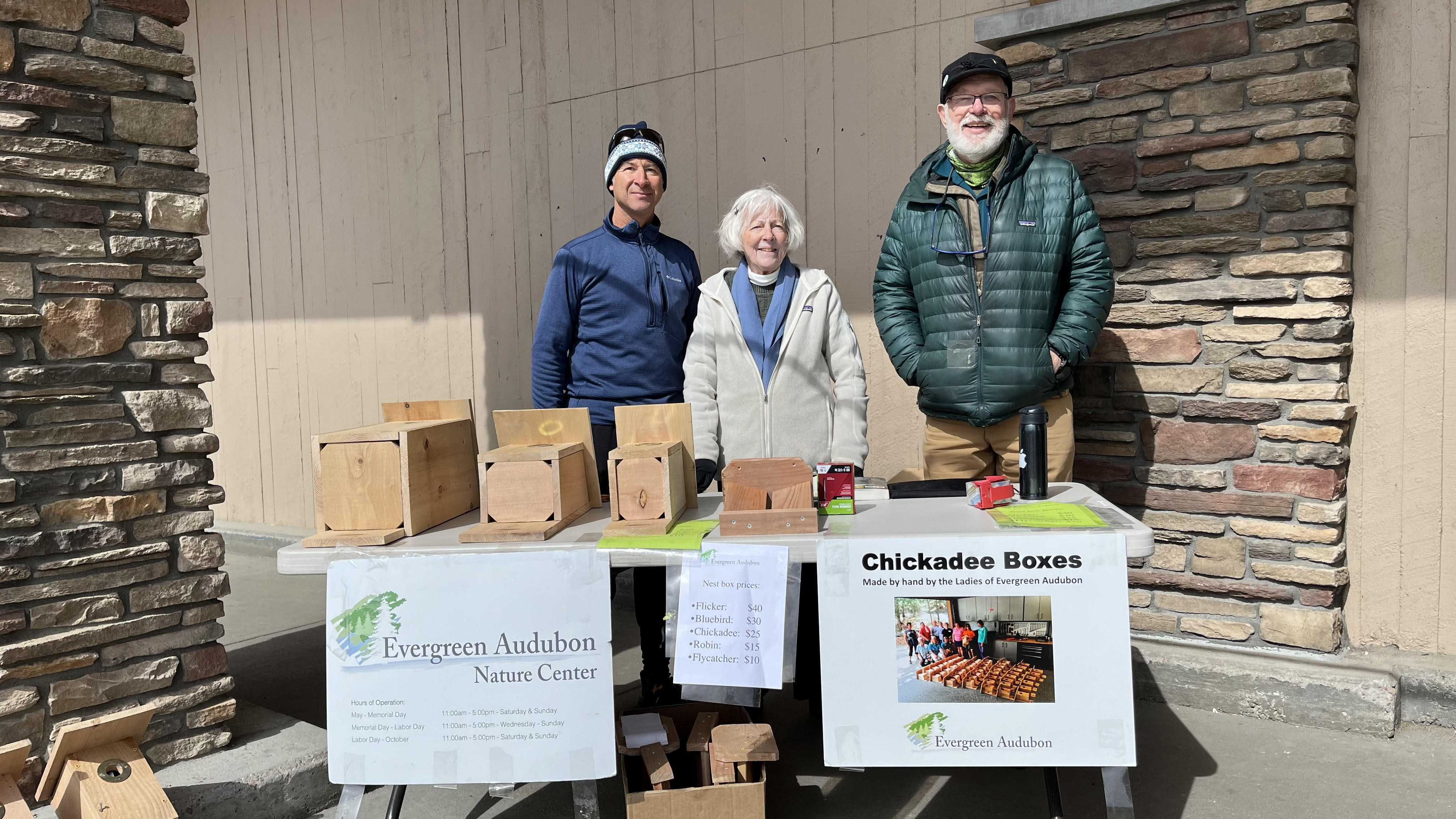 Two men and a women standing behind a table of nest boxes (birdhouses).