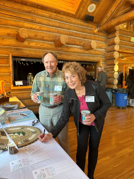 A smiling man holding cups in each hand. A woman holding a cup and raising her silent auction bid. The bid sheet is on a table covered with a white table cloth.