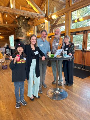 A young boy drinking a Coke, a woman, two men then another woman, all standing at a stand-up cocktail table and laughing. Behind them are windows with sun streaming through and rafters hung with decorative lights.