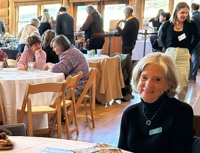 A blonde woman smiling sitting at a table and smiling at the camera. There are other tables behind her with people at them, a line of people behind the tables, and windows in the background.