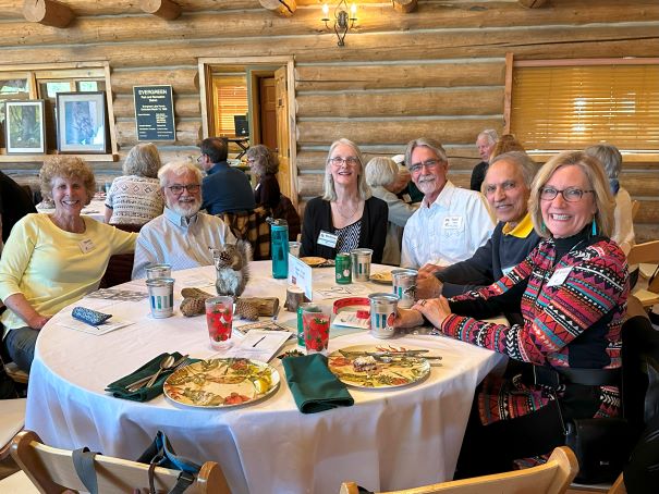 Three couples sitting at a table, smiling at the camera. Behind them are other people sitting at tables and a log wall.