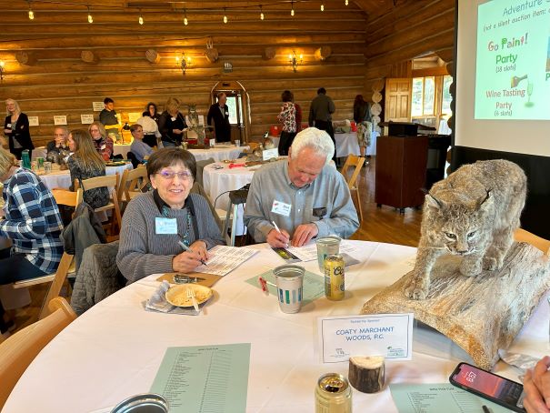 A man and a woman sitting at a table. They are filling out raffle tickets. There is a taxidermy bob cat on the talbe and you can see other tables and a screen with a slide show behind them.