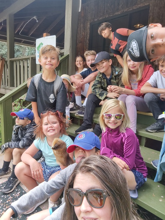 Several brightly dressed youngsters and a woman who is wearing dark glasses, pose on the porch steps of a old looking cabin. Everyone is either smiling or making funny faces. One girl is holding a fuzzy brown puppet, two of the boys are wearing brigth blue Chicago Cubs hats, one boy is standing and making a silly face while another boy is completely sideways in the photo.