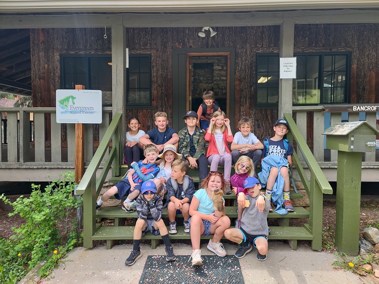 Several brightly dressed youngsters sitting on the green wooden steps of an old, wooden cabin. There is a mailbox on a post on one side and a sigh that says, Evergreen Nature Center on the other side, along with a green bush and some yellow dandelions. You can see two windows and the front door of the cabin behind the children.