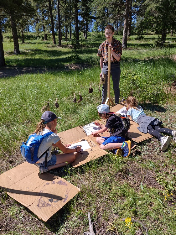 Three children sitting on the ground and working on art projects. There is an adult watching and the setting is a grassy spot among several trees.