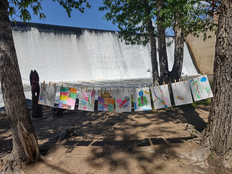 Several colorful watercolor paintings are displayed on a string strung between two trees. There is a dam and waterfall in the background.