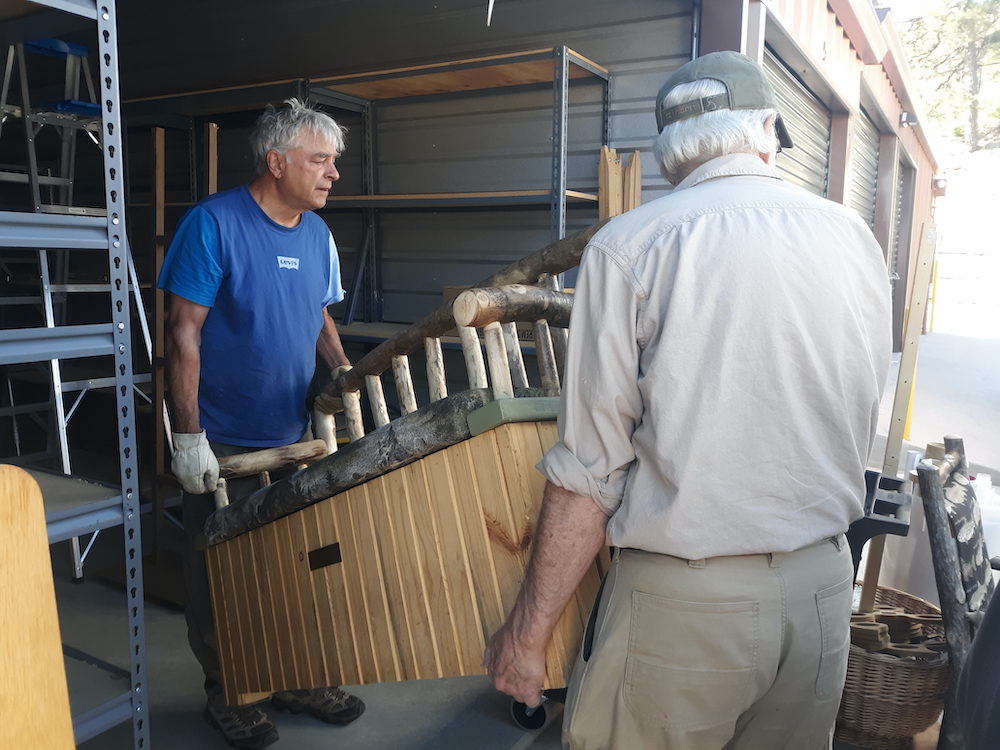 Two white haired men, one in blue the other in khakis, carry a wooden and upholstery bench out of a storage until lined with metal shelves and other miscellaney.