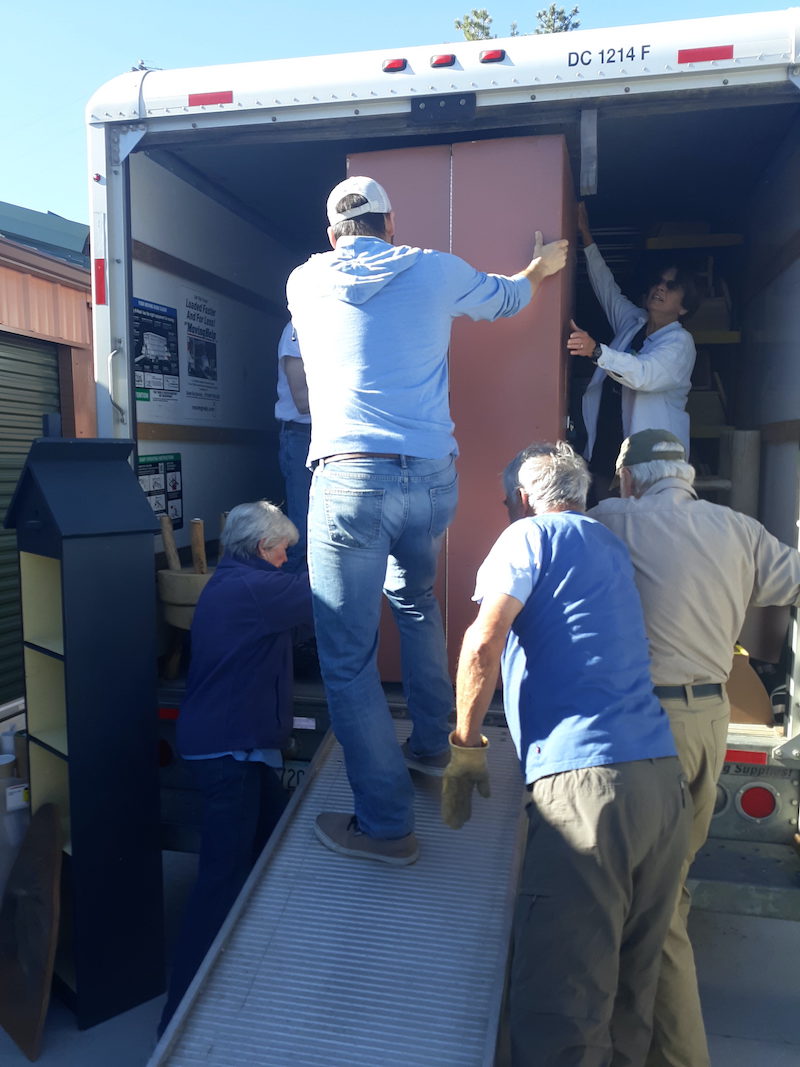Six people, one on a ramp, moving a tall cabinet that barely fits, into the back of a truck. There are a few other pieces of furniture in the picture as well.