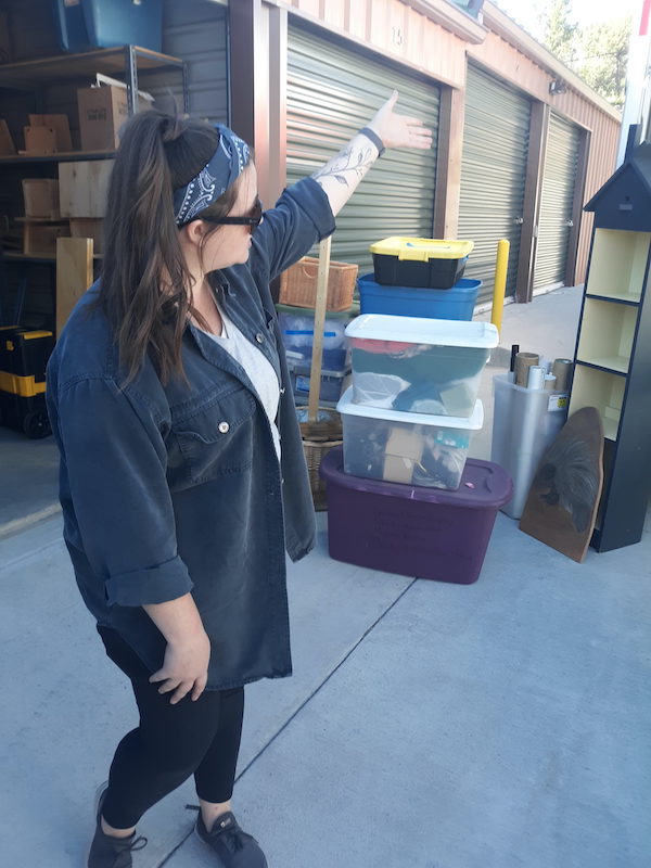 A woman, dressed in black shoes and slacks, a white Tshirt with a black overshirt, long brown hair held back with a blue hair band, is gesturing with her left hand towards something out of the photo frame. There are bins and boxes as well as miscellaneous pieces of furniture behind her. You can also see several other storage units in the background.