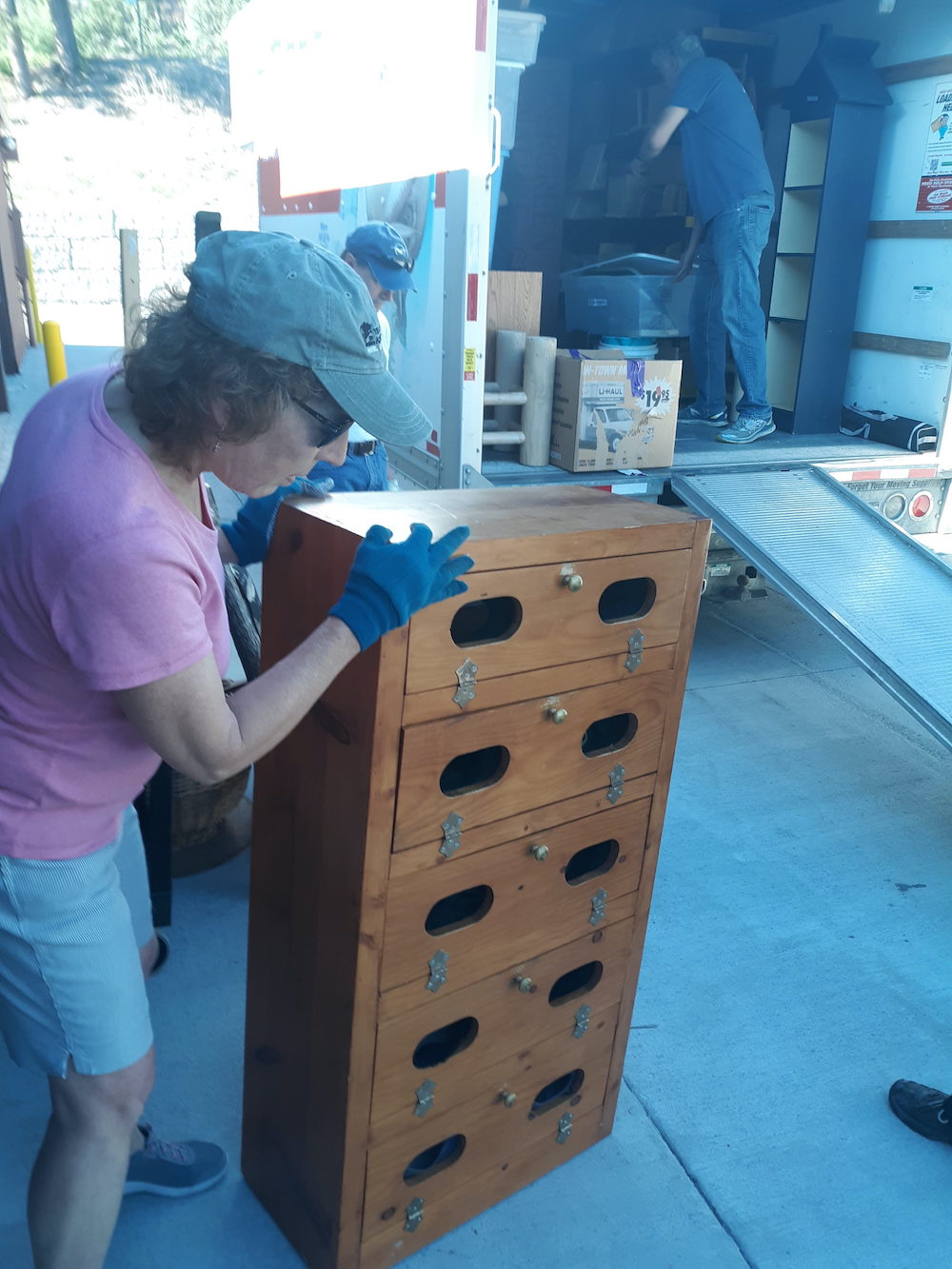 A woman with curly hair, wearing a baseball cap, a pink Tshirt, white shorts and blue shoes, moving a wooden cabinet with hinged drawers, towards the back of an open truck. There are moving boxes and other items in the truck, and two men in the picture, one next to the woman and one in the truck. There is also a ramp leading up to the truck's floor from the ground.