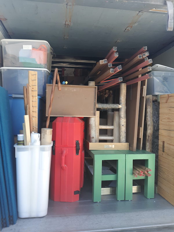 Open back of a truck showing bright blue panels, white and blue plastic bins, a bright orange container, green tables, a wood and upholstered bench and several other odd and ends. The truck is tightly packed.