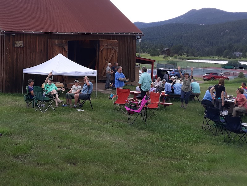 Several colorfully dressed people sitting in groups on camp chairs and picnic tables set on green grassi. There is a barn, some parked cars, a pond, and tree covered hills in the background.