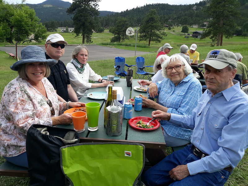 Six smiling people sitting at a dark green picnic table. From left to right: A woman in a printed blouse wearing a blue hat; a man in a black shirt, dark glasses and a white hat; a woman in a white shirt; a woman with white hair wearing dark glasses, a women with white hair wearing a blue plaid shirt and a man wearing a long-sleeved blue shirt, jeans and a green hat. IN the background are more people, two blue camp chairs, a basketball court, a green lawn, trees and a hill.