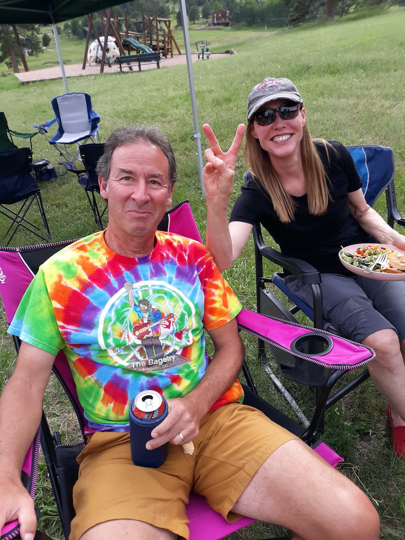 A man in a brightly colored tie-dyed T-shirt, a woman with long brown hair wearing dark glasses and a black t-shirt, holding a plate of food. Both are smiling broadly and the woman is flashing a peace sign.