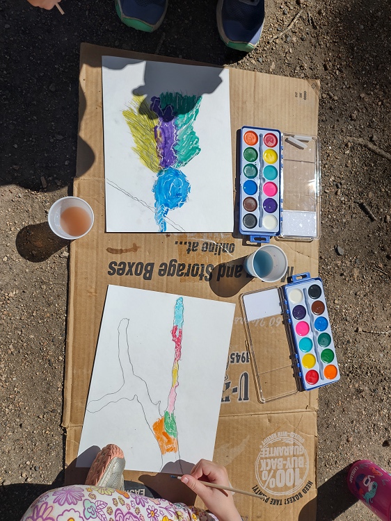 overhead photo of children (you can only see their hands and feet) painting nature scenes with watercolor. The watercolor paints are off to the side.