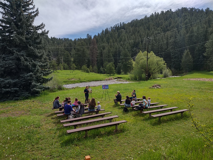 A green meadow with a stream running through it, a big pine tree on one side and a forested hillside behind. The meadow rows of wooden benches. There are families sitting on the benches facing an adult story teller.