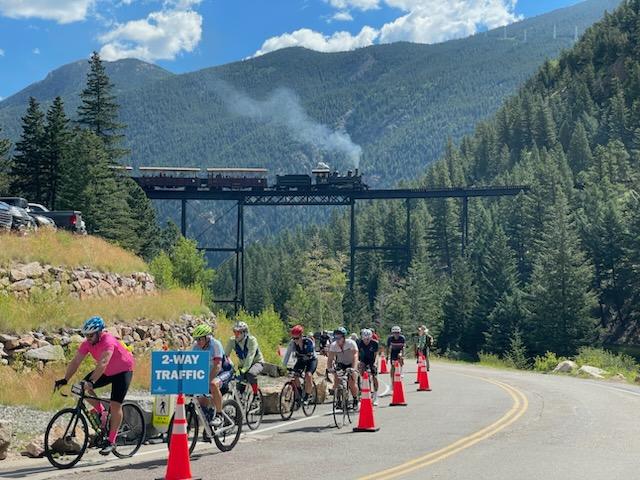 A group of colorfully dressed bicyclists riding up a hill. There are traffic cones and a sign that says "2-way traffic." There is a train high up on a trestle bridge in the background and tree covered mountains.