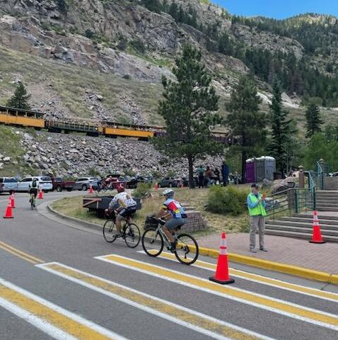 Two bicyclists on the road, a yellow cross walk, orange traffice cones, a woman in a safety vest on the sidewalk, cars parked in the back and an old fashioned tourist train passing by on a stone embankment.