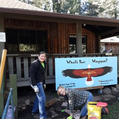 two women, one standing the other kneeling installing paver in front of a wooden sign with the wingspans of an eagle, hawk, kestrel, and a hummingbird