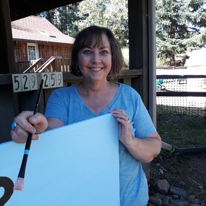 smiling women behind a wooden sign holding a paint brush