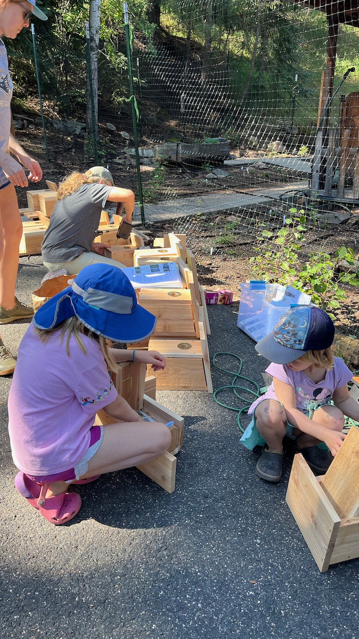 two children with nestboxes on the ground outside, with a person in the background drilling on a nestbox