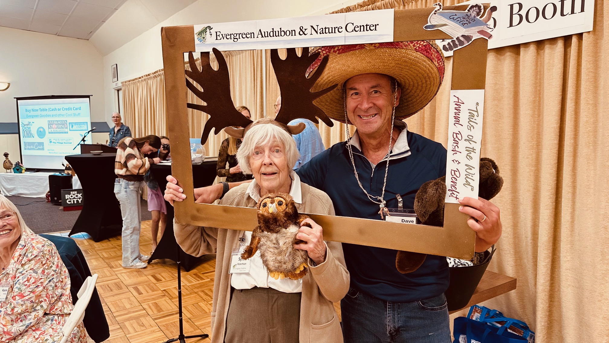 two people with silly hats, holding stuffed animals taking a selfie with a frame around them
