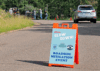 A large orange sign in the middle of the road warning, "Please, slow down, roadside mitigation event" in orange, white, and blue letters. It's against a backdrop of workers cutting down trees in a forest along a road.