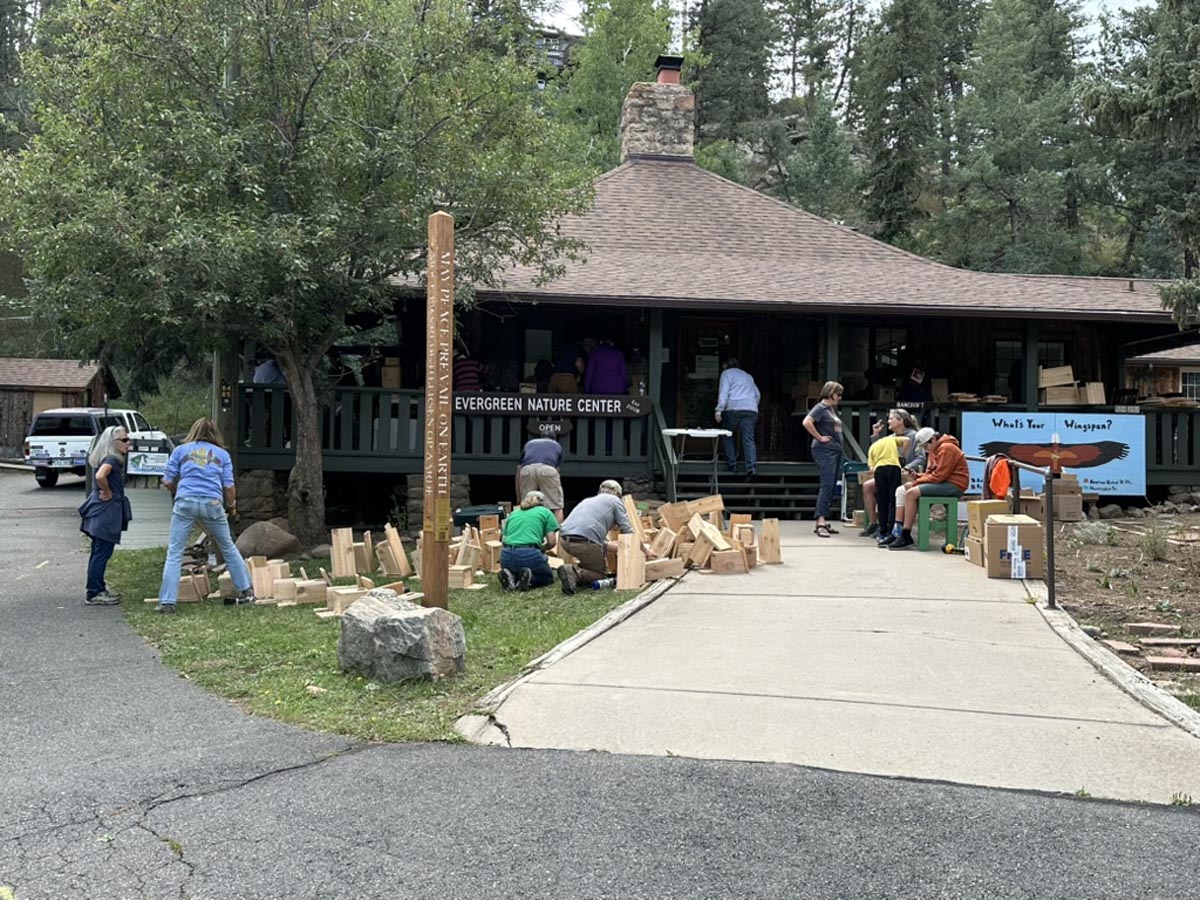 A view from the parking lot of the nestbox building scene with completed nestboxes piled on the front lawn and people scattered around doing different tasks.