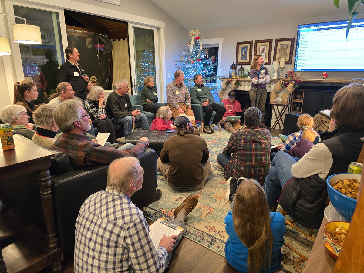 a bunch of people in a family room sitting on the floor and in chairs looking at a screen over the fireplace
