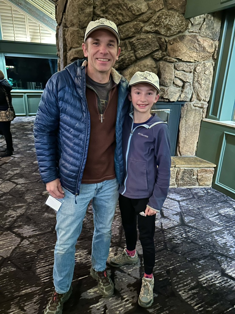 Man on left, little girl on right, standing on a carpet in front of a fireplace.