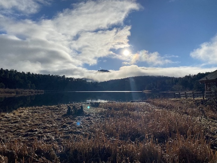 The sun shining through the clouds in a blue sky over a marshy area by a lake.