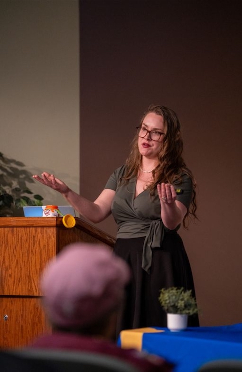 A woman with curly red hair in a formal grey top and black skirt, presenting in front of an audience, using her hands to talk next to a wooden podium.